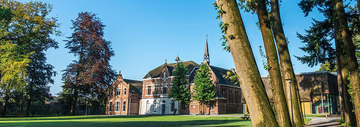 zicht op het klooster vanaf het pad dat naar de ingang leidt, een grasveld met een oud gebouw en hoge oude bomen
